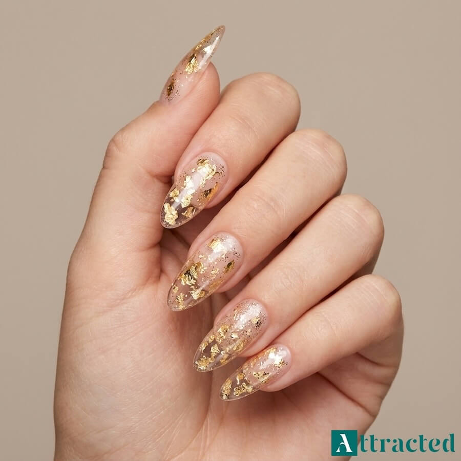 Zbliżenie jednej dłoni z długimi migdałkowymi paznokciami konstrukcyjnymi z przezroczystą końcówką wypełnioną złotą folią i brokatem, na tle jasnego studia