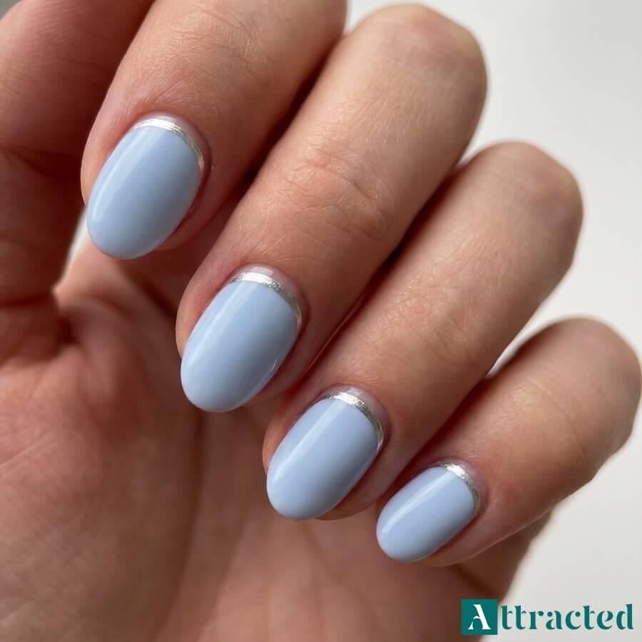 Zbliżenie jednej dłoni z krótkimi owalnymi paznokciami w pastelowym błękicie i srebrną linią reverse french, na jasnym neutralnym tle