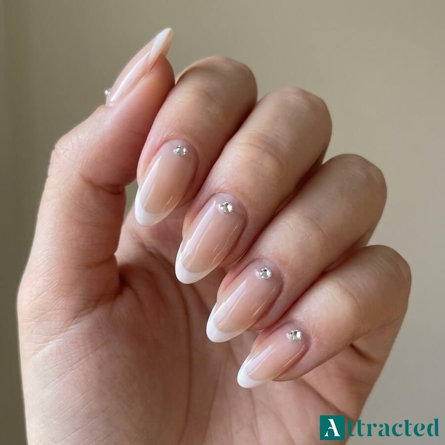 Zbliżenie jednej dłoni z migdałkowymi paznokciami french i pojedynczym kryształkiem przy nasadzie każdego paznokcia, na tle jasnym i neutralnym